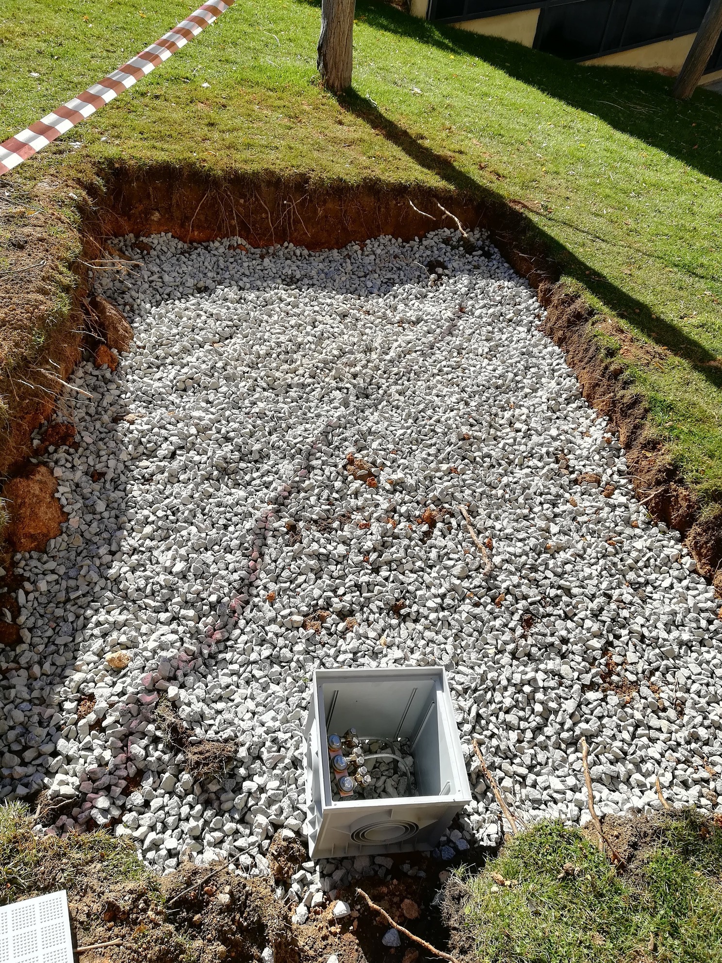 Zanja con grava y caja de conexiones plásticas en área verde, rodeada de tierra y cinta de seguridad al fondo.