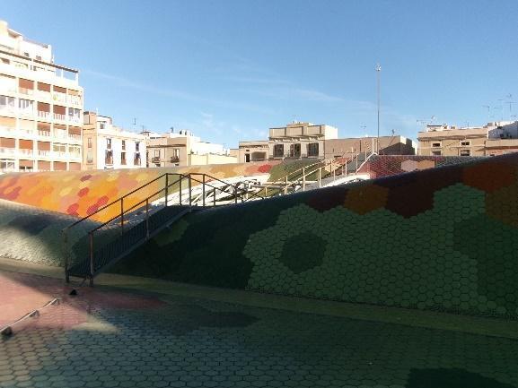 Una vista desde el nivel de la calle o un punto más bajo, mostrando las ondulaciones del tejado de mosaicos del Mercado de Santa Caterina y una pasarela de acceso, con edificios urbanos al fondo bajo un cielo azul.