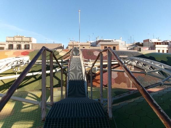 Una pasarela de rejilla metálica con barandillas oxidadas, que atraviesa el tejado ondulado y multicolor del Mercado de Santa Caterina, con vistas a los edificios circundantes bajo un cielo azul despejado.
