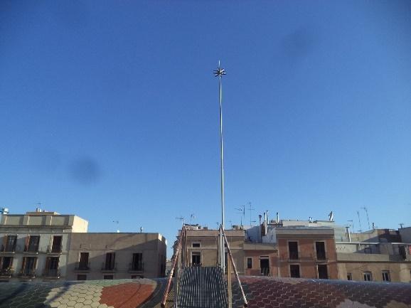 Un mástil metálico alto con un remate ornamentado, erigiéndose sobre el colorido tejado de mosaicos del Mercado de Santa Caterina, con edificios urbanos al fondo bajo un cielo azul claro.