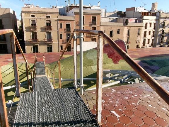 Una pasarela metálica y barandillas oxidada en primer plano, conduciendo a una azotea con tejado de mosaicos coloridos, y edificios residenciales típicos de Barcelona al fondo.