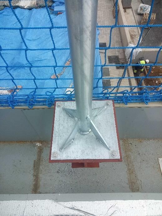 Primer plano de la robusta base de un mástil o poste metálico, fijado a una superficie, con una red de seguridad azul visible al fondo.