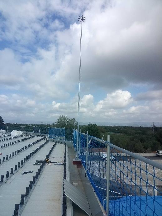 Vista de una amplia azotea con hileras de soportes y un mástil alto, protegida por barandillas con red de seguridad azul, bajo un cielo parcialmente nublado.