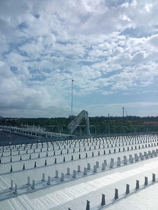 Una vista de una azotea plana con numerosas hileras de soportes o fijaciones blancas y negras, una estructura mecánica angular en el centro y un mástil delgado, todo bajo un cielo parcialmente nublado.