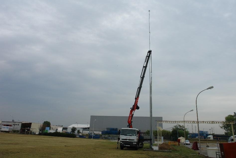 Una grúa montada sobre camión elevando una sección de una torre alta en un área de terreno llano, con edificios industriales y otra infraestructura al fondo, bajo un cielo nublado.