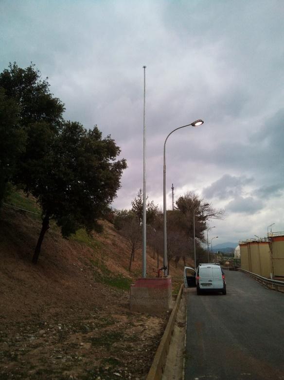 Vista de una farola moderna iluminando una carretera adyacente a una ladera con vegetación, con estructuras industriales al fondo bajo un cielo nublado.