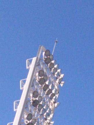 Detalle de la sección superior de una torre de iluminación de estadio, mostrando una densa disposición de focos de gran potencia y un pararrayos en la cima, contra un cielo despejado.
