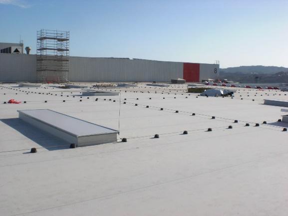 Amplia azotea plana de un edificio industrial o comercial, equipada con un sistema de línea de vida para seguridad, un tragaluz alargado y andamios, indicando actividades de construcción o mantenimiento.