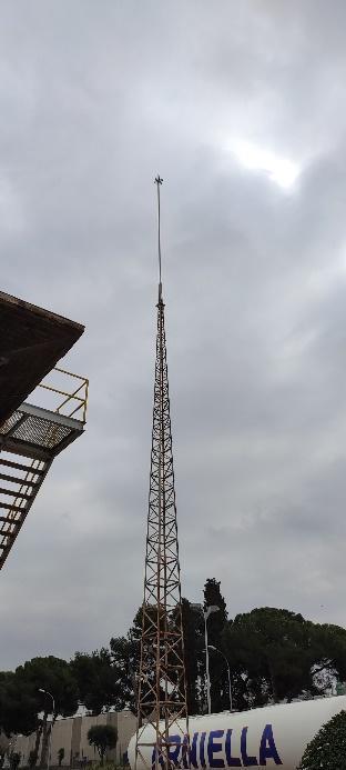 Una alta torre de celosía, probablemente para comunicaciones o antenas, junto a una escalera industrial y un tanque, bajo un cielo nublado con un sol velado.