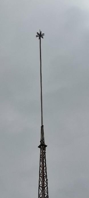 Primer plano de la aguja superior de una torre metálica, probablemente una antena o un pararrayos, con una estructura puntiaguda en su extremo, contra un cielo gris uniforme.