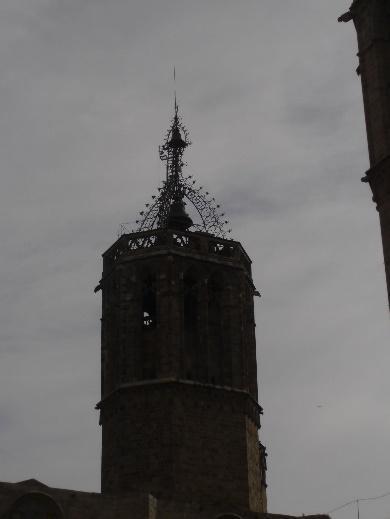 Primer plano de la parte superior de un antiguo campanario de piedra con detalles góticos y un chapitel calado, destacando contra un cielo gris y cubierto.