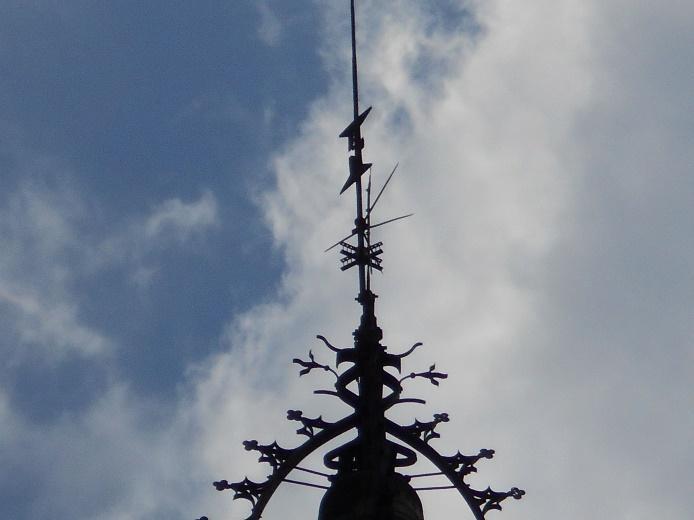 Primer plano de la parte superior de un chapitel ornamentado de una torre, con detalles de hierro forjado y una aguja vertical, silueteados contra un cielo parcialmente nublado.