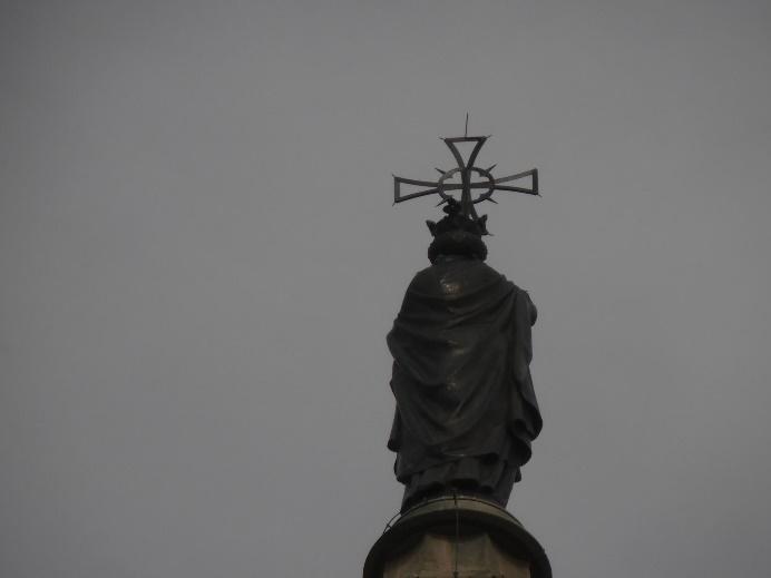 Vista trasera de una estatua monumental de figura envuelta en ropajes, coronada por una cruz ornamentada, silueteada contra un cielo completamente cubierto.