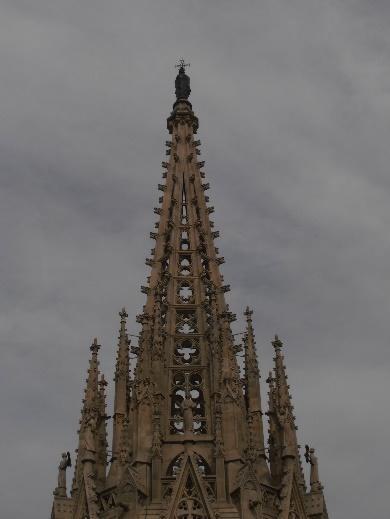 Vista ascendente de la imponente aguja central de una catedral gótica, detallada con pináculos y elementos arquitectónicos, contra un cielo cubierto.