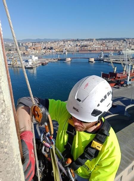 Un técnico especializado en acceso con cuerda inspeccionando su equipo con vistas a un puerto marítimo.