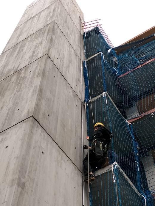 Un trabajador con casco y arnés desciende por la fachada de un edificio en construcción con una red de seguridad azul.