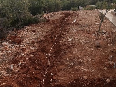 Zanja excavada: La línea visible en el suelo que ha sido removida, indicando el lugar donde se está instalando el sistema.
Cable o tubería (línea clara): El elemento principal que se está tendiendo a lo largo de la zanja. Podría ser un cable conductor de tierra o una tubería.
Terreno rocoso/arcilloso: El suelo de color rojizo con presencia de piedras, característico de zonas con este tipo de geología.
Vegetación de monte bajo y árboles: La flora que se observa en los alrededores, indicando un paisaje natural o semi-natural.
Horizonte/Paisaje de fondo: Las colinas o montañas en la distancia, sugiriendo una ubicación rural o de campo.