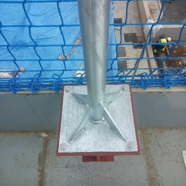 Primer plano de la robusta base de un mástil o poste metálico, fijado a una superficie, con una red de seguridad azul visible al fondo.