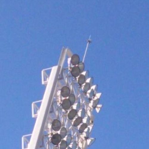 Detalle de la sección superior de una torre de iluminación de estadio, mostrando una densa disposición de focos de gran potencia y un pararrayos en la cima, contra un cielo despejado.