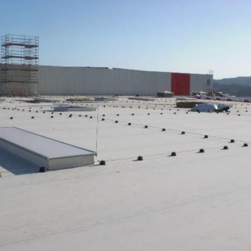 Amplia azotea plana de un edificio industrial o comercial, equipada con un sistema de línea de vida para seguridad, un tragaluz alargado y andamios, indicando actividades de construcción o mantenimiento.