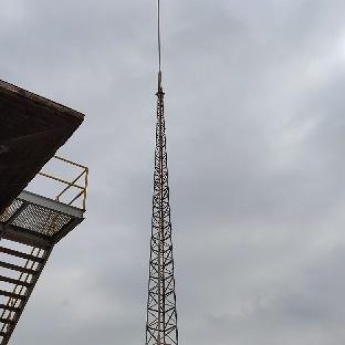 Una alta torre de celosía, probablemente para comunicaciones o antenas, junto a una escalera industrial y un tanque, bajo un cielo nublado con un sol velado.