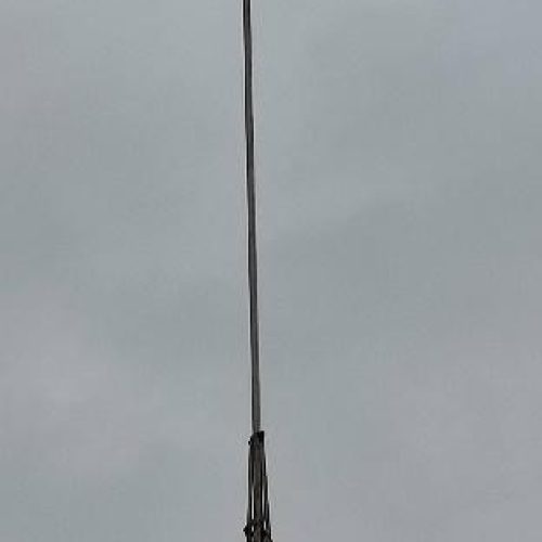 Primer plano de la aguja superior de una torre metálica, probablemente una antena o un pararrayos, con una estructura puntiaguda en su extremo, contra un cielo gris uniforme.