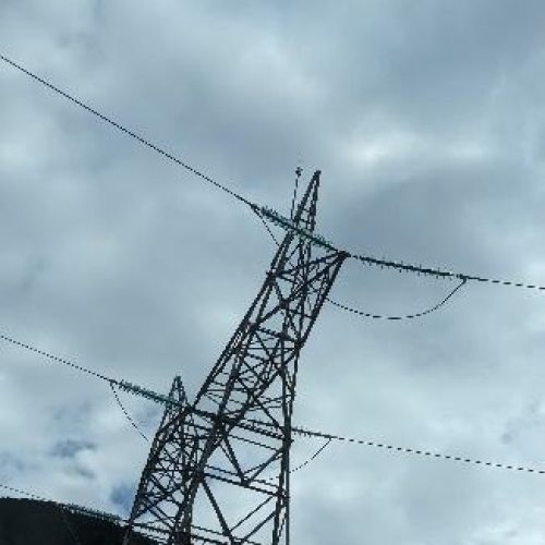 Vista desde abajo de una torre de tendido eléctrico de alta tensión, con sus cables y aisladores, silueteada contra un cielo con nubes dispersas.