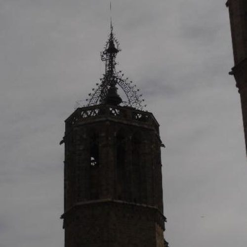 Primer plano de la parte superior de un antiguo campanario de piedra con detalles góticos y un chapitel calado, destacando contra un cielo gris y cubierto.