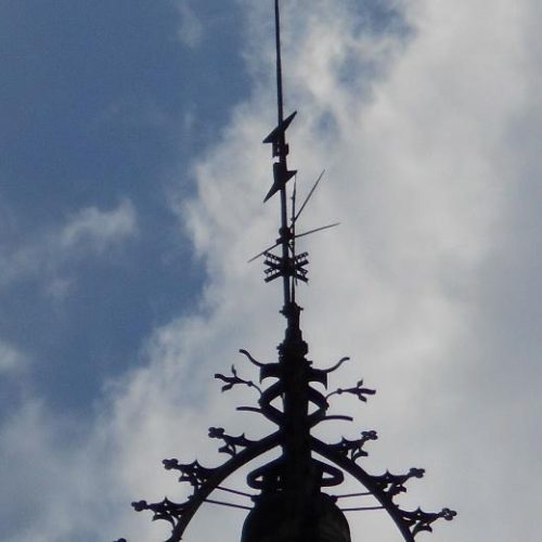 Primer plano de la parte superior de un chapitel ornamentado de una torre, con detalles de hierro forjado y una aguja vertical, silueteados contra un cielo parcialmente nublado.