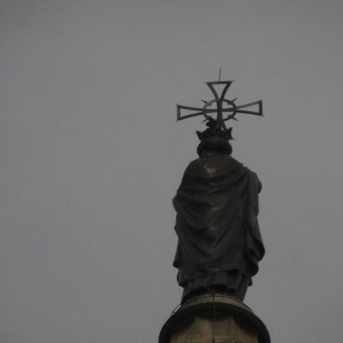 Vista trasera de una estatua monumental de figura envuelta en ropajes, coronada por una cruz ornamentada, silueteada contra un cielo completamente cubierto.