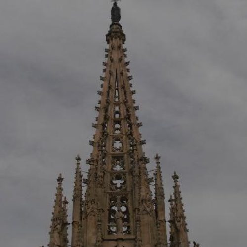 Vista ascendente de la imponente aguja central de una catedral gótica, detallada con pináculos y elementos arquitectónicos, contra un cielo cubierto.