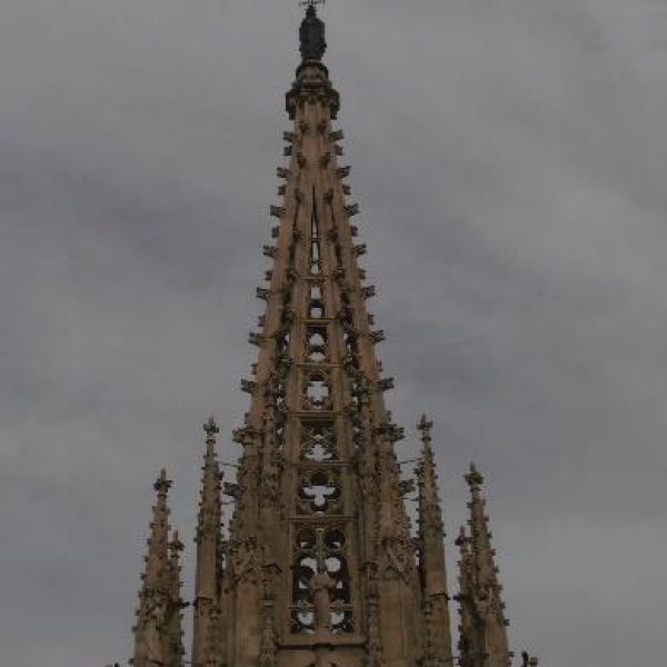 Vista ascendente de la imponente aguja central de una catedral gótica, detallada con pináculos y elementos arquitectónicos, contra un cielo cubierto.