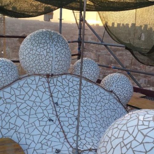 Primer plano de las esferas y superficies curvas cubiertas de trencadís en una de las torres de la Sagrada Familia, con el horizonte de la ciudad visible al fondo.