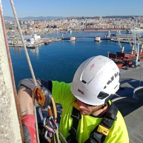 Un técnico especializado en acceso con cuerda inspeccionando su equipo con vistas a un puerto marítimo.