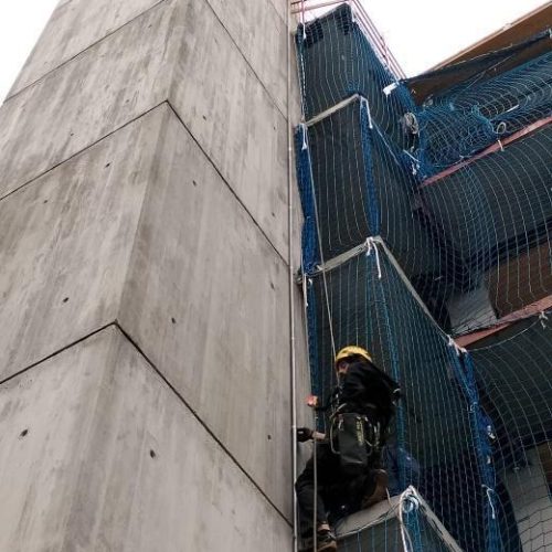 Un trabajador con casco y arnés desciende por la fachada de un edificio en construcción con una red de seguridad azul.
